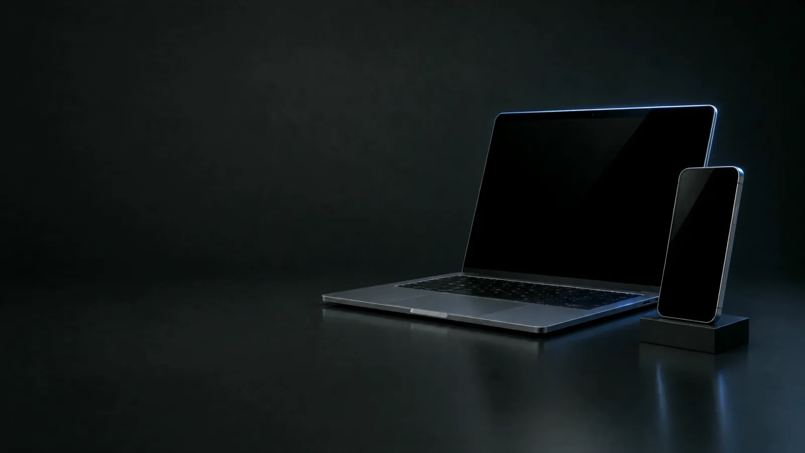 A laptop and a phone on a dark desk, lit by a soft blue glow